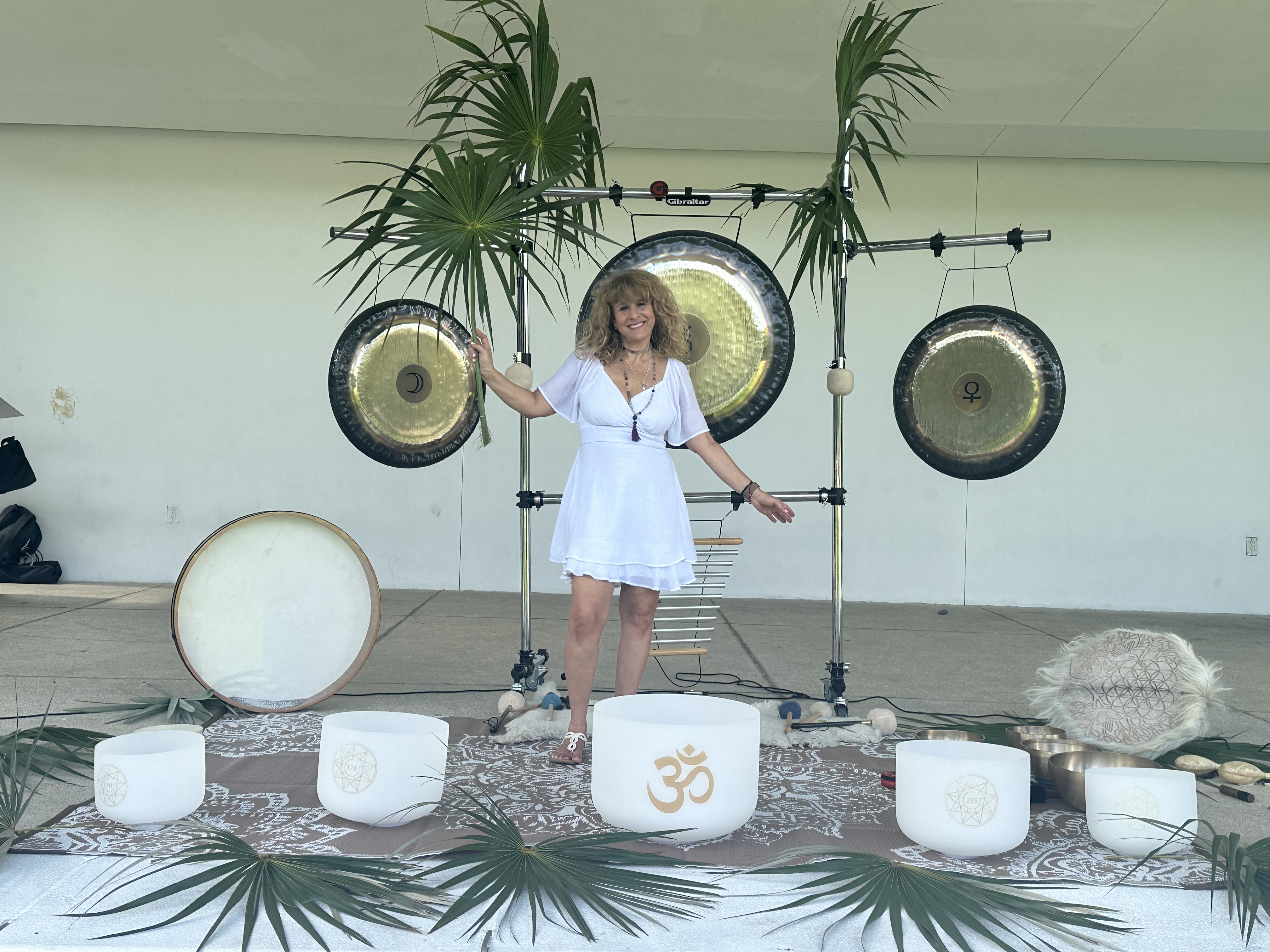 Diana smiling with three planetary gongs, frosted crystal singing bowls, a sacred drum, and palm fronds