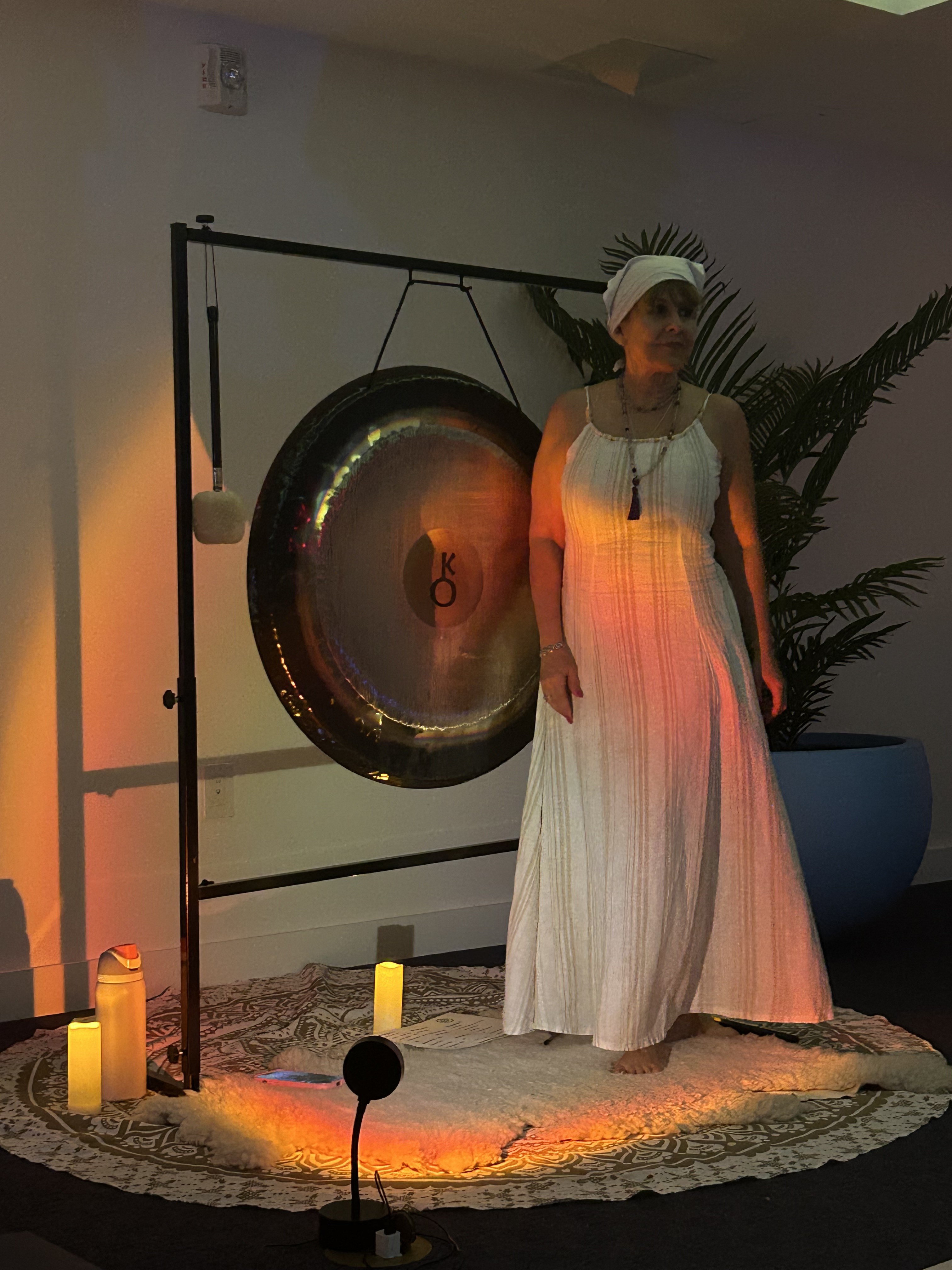 Diana standing in a white dress and headwrap beside the Chiron gong in warm candlelight