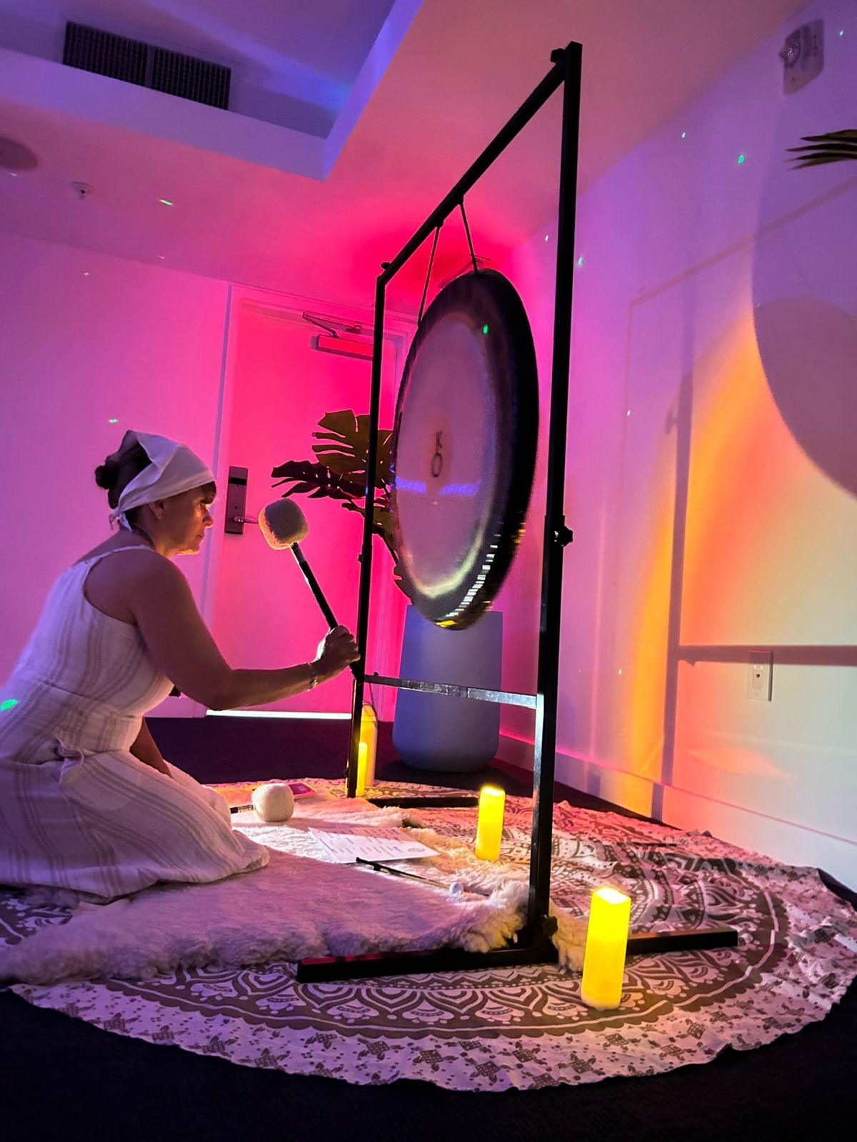 Diana kneeling in white while playing the Chiron gong among warm candles and magenta ambient light