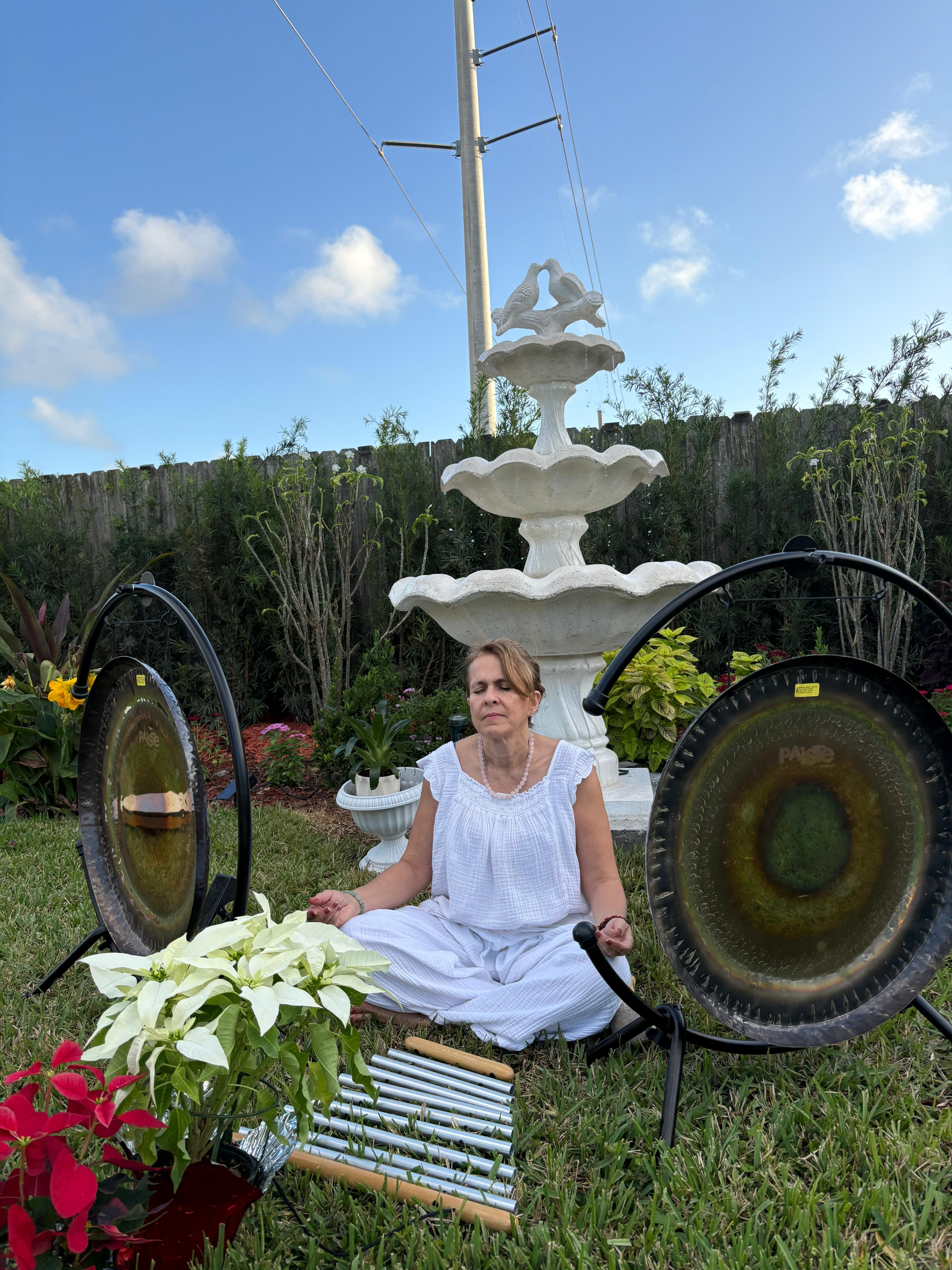Diana meditating in a garden between two Paiste gongs beside a tiered fountain and bar chimes