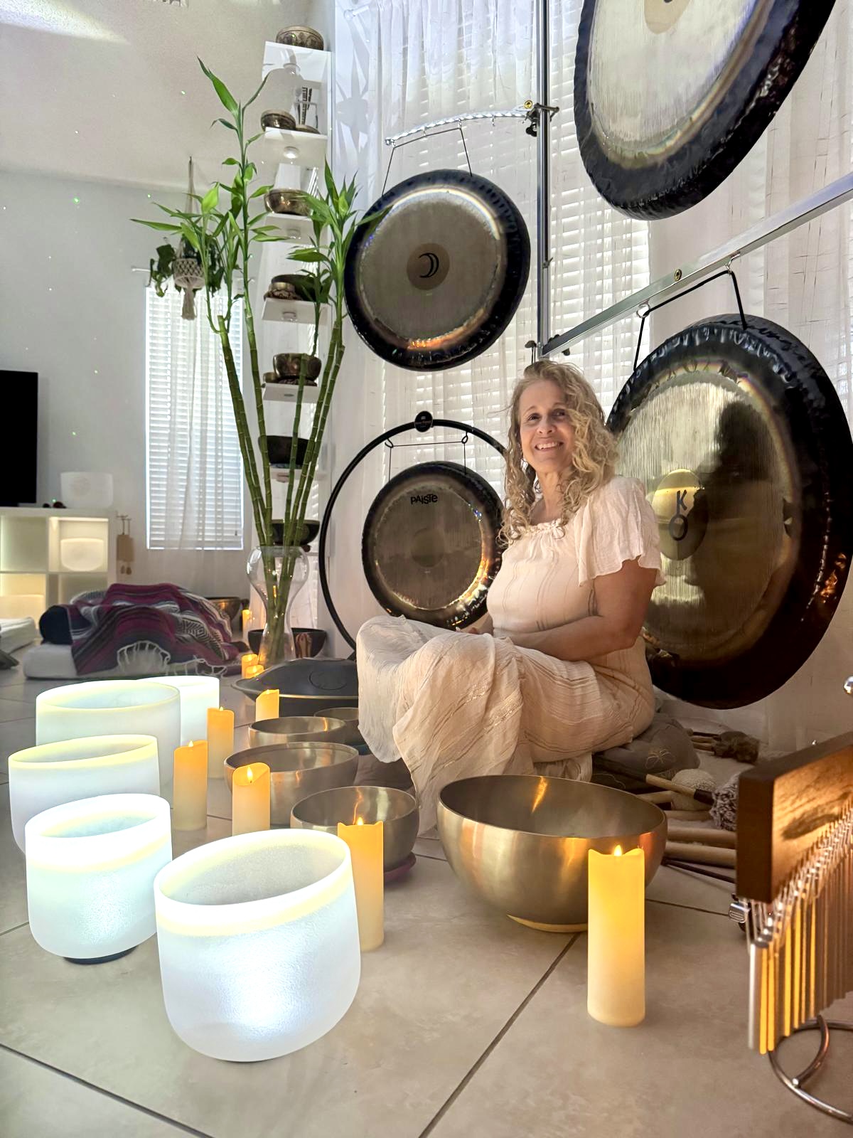 Diana Sawal seated in white among glowing crystal bowls, Tibetan bowls, candles, and hanging gongs